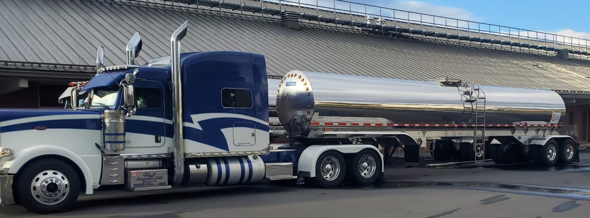 A Wall Transportation blue Peterbilt coupled to a polished stainless food-grade tank trailer, full side profile at a processing facility.
